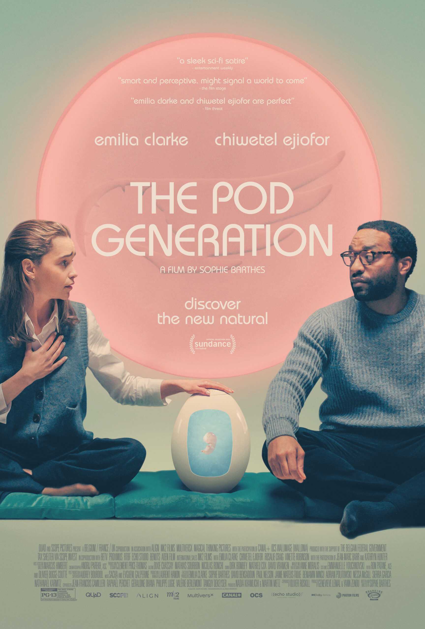 Emilia Clarke and Chiwetel Ejiofor sit legs crossed with a white pod between them, in which a fetus can be seen.