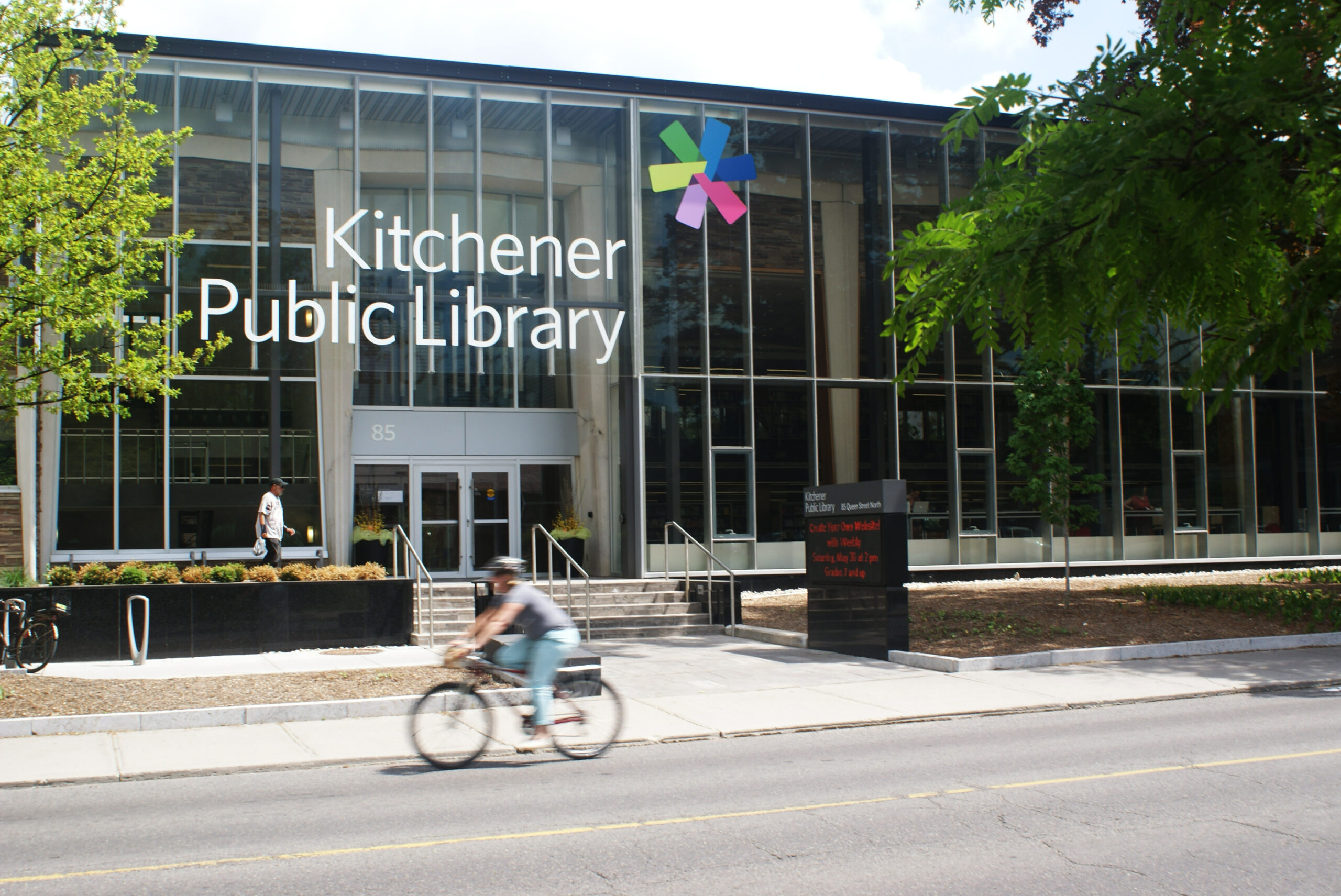 The front doors of the KPL Central Branch, whose front wall is entirely windows. A cyclist rides on the road in front.